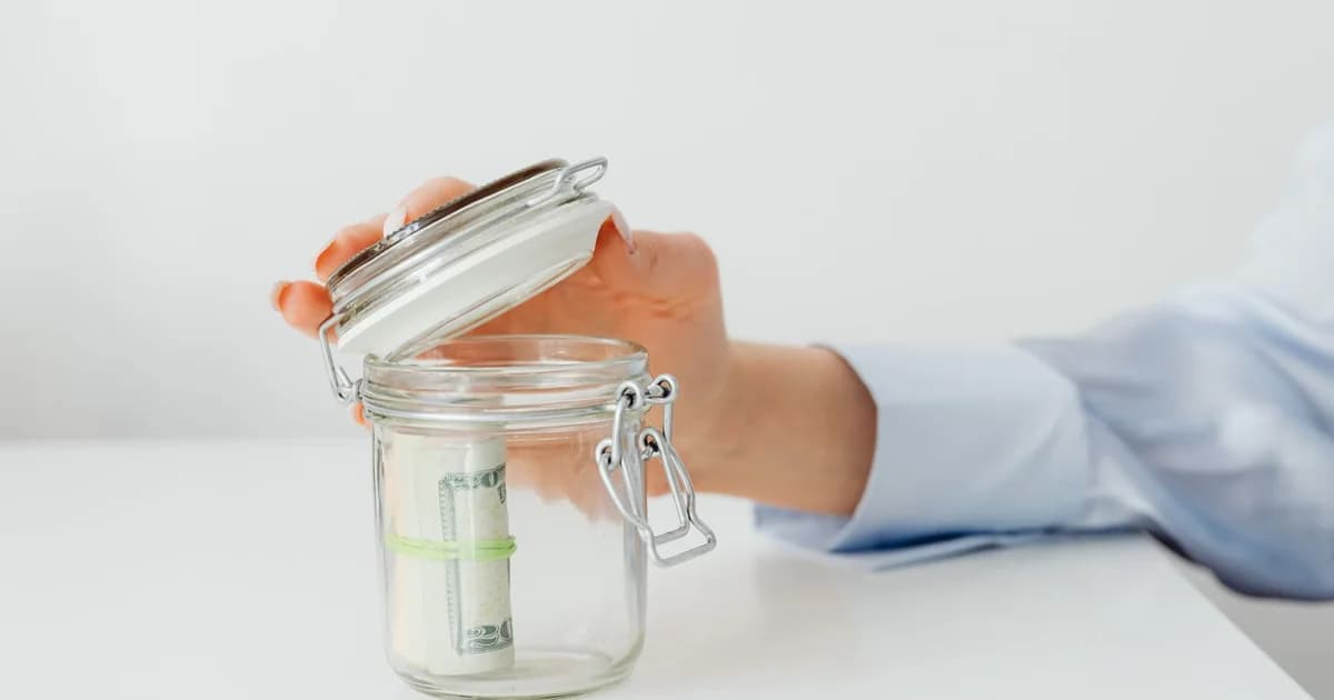 A hand places money in a glass jar on a white table, symbolizing savings.