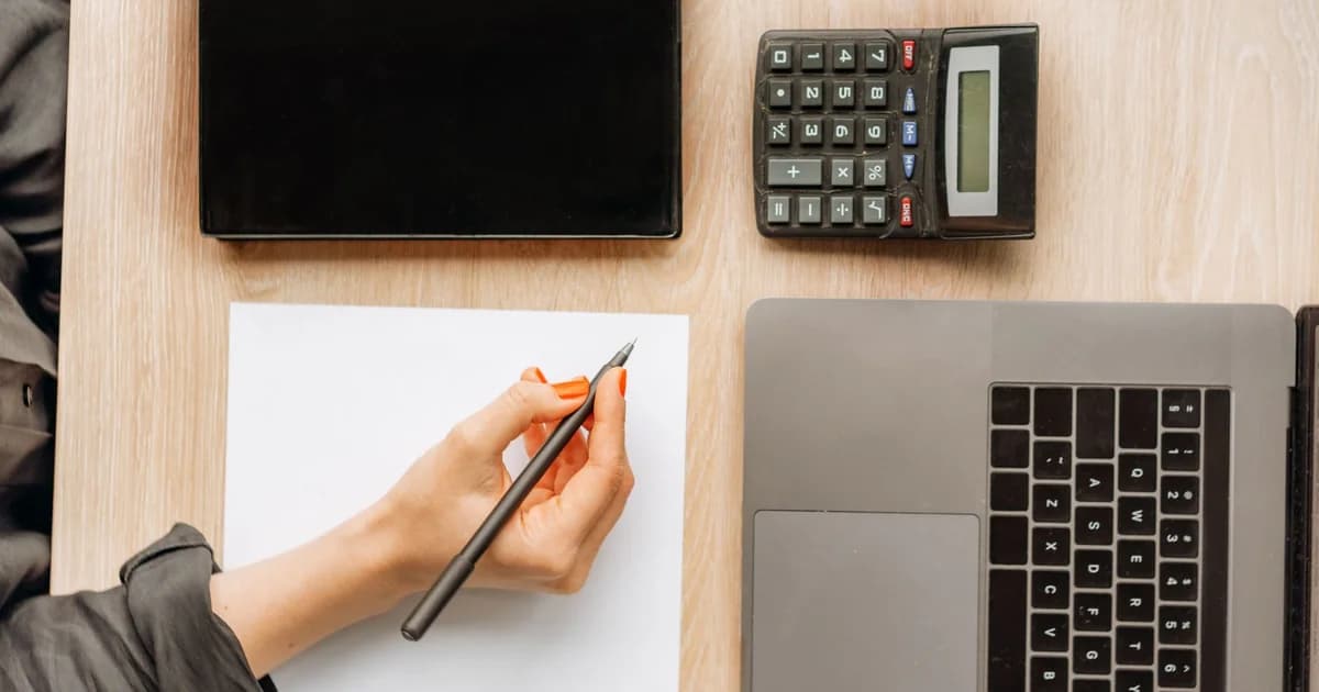 A person writing on paper surrounded by a laptop, tablet, and calculator on an office desk.